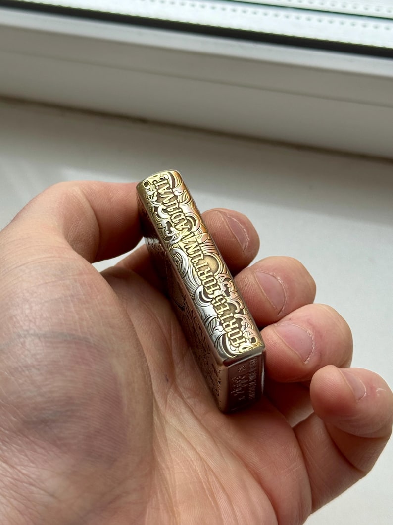May include: A rectangular, metallic lighter with ornate engravings. The lighter has a gold and silver finish, with the text "FORTES FORTUNA ADIUVAT" etched along the side. The lighter is held in a hand.