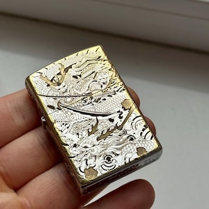 Può includere: Un accendino rettangolare in metallo con una dettagliata incisione color oro e argento. Il disegno raffigura un samurai in armatura tradizionale che impugna una katana sopra un grande drago, con elementi architettonici e motivi floreali.