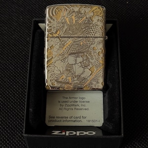May include: A silver-tone metal lighter featuring an intricate engraved design of a samurai warrior and a dragon with gold-colored accents. The lighter is presented in a black box with a card stating: The Armor logo is used under license by ZippMark, Inc. All Rights Reserved. See reverse of card for product information. 181501-I Zippo.