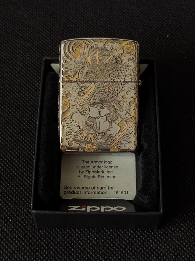 May include: A silver-tone metal lighter featuring an intricate engraved design of a samurai warrior and a dragon with gold-colored accents. The lighter is presented in a black box with a card stating: The Armor logo is used under license by ZippMark, Inc. All Rights Reserved. See reverse of card for product information. 181501-I Zippo.
