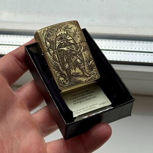 May include: A brass-colored lighter with an engraved design of a wolf-like figure in a forest scene. The lighter is in a black box, and a small card with text is visible. The text on the card reads "The Armor logo is used under license by ZippMark, Inc. All Rights Reserved."