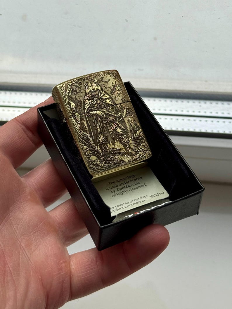 May include: A brass-colored lighter with an engraved design of a wolf-like figure in a forest scene. The lighter is in a black box, and a small card with text is visible. The text on the card reads "The Armor logo is used under license by ZippMark, Inc. All Rights Reserved."