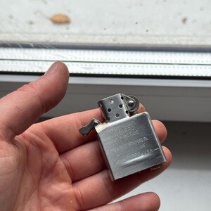 May include: A silver metal Zippo lighter with text on the side. The lighter is held in a hand. The text on the lighter reads "Zippo Bradford, PA. Made in U.S.A."