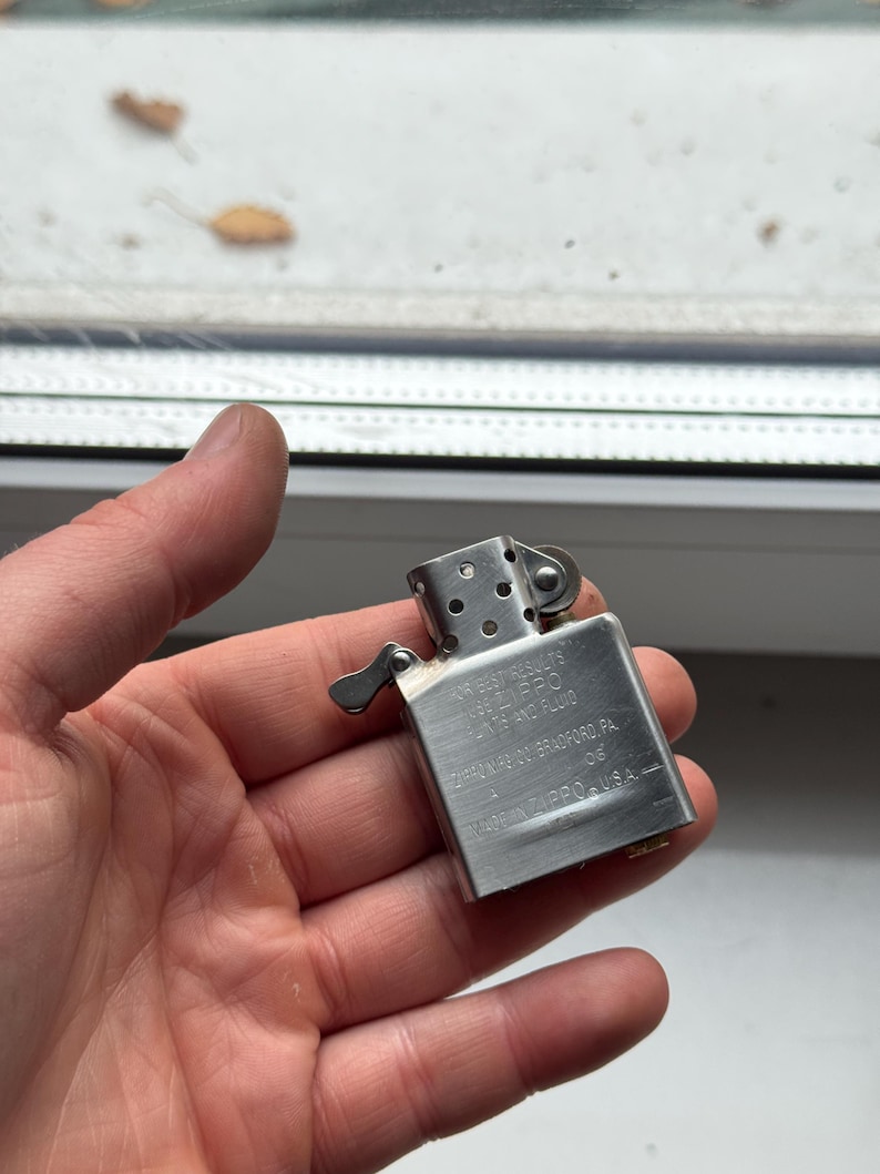 May include: A silver metal Zippo lighter with text on the side. The lighter is held in a hand. The text on the lighter reads "Zippo Bradford, PA. Made in U.S.A."