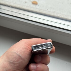 May include: A silver Zippo lighter held in a hand. The lighter is rectangular with rounded edges and has the Zippo logo and text "BRADFORD PA. MADE IN U.S.A." engraved on it. The lighter is closed and appears to be in good condition.