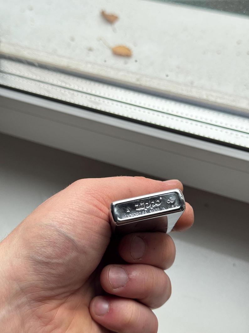 May include: A silver Zippo lighter held in a hand. The lighter is rectangular with rounded edges and has the Zippo logo and text "BRADFORD PA. MADE IN U.S.A." engraved on it. The lighter is closed and appears to be in good condition.