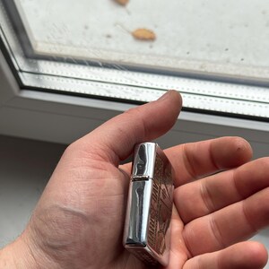 May include: A silver metal lighter with an engraved design. The lighter is held in a hand and has a rectangular shape with rounded edges. The engraved design appears to be a logo or emblem.