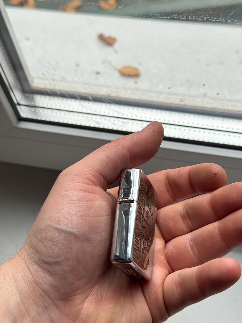 May include: A silver metal lighter with an engraved design. The lighter is held in a hand and has a rectangular shape with rounded edges. The engraved design appears to be a logo or emblem.