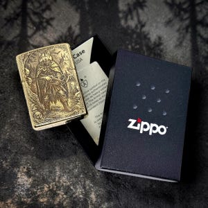 May include: A gold-colored Zippo lighter with an engraved design of a warrior figure and wolf head. The lighter is open, next to its black box with the Zippo logo. The box also contains a white insert with text.