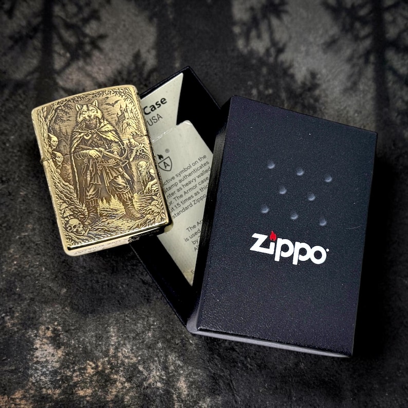 May include: A gold-colored Zippo lighter with an engraved design of a warrior figure and wolf head. The lighter is open, next to its black box with the Zippo logo. The box also contains a white insert with text.