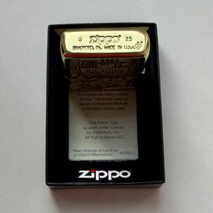Pu&ograve; includere: Un accendino Zippo color oro in una scatola nera. L'accendino &egrave; contrassegnato con "Zippo 25 BRADFORD, PA. MADE IN U.S.A." e include una grafica. La scatola mostra anche il logo Zippo e le informazioni sul prodotto.