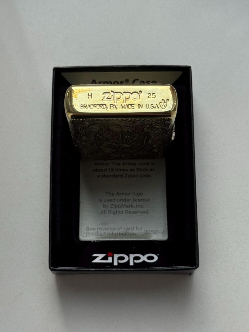 May include: A gold-colored Zippo lighter in its original black box. The lighter is engraved with a detailed pattern and the top is marked with "Zippo 25 BRADFORD, PA, MADE IN U.S.A." The box includes a card with information about the Armor case.