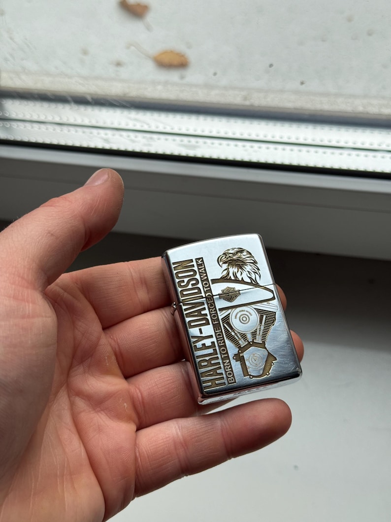 May include: A silver Harley-Davidson lighter with gold-colored accents. The lighter features an eagle graphic and the text "HARLEY-DAVIDSON BORN TO RIDE FORCED TO WALK". The design includes an engine illustration.