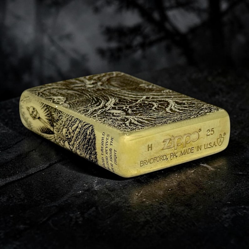 May include: A brass-colored Zippo lighter with an engraved design. The lighter features intricate artwork and the text "AND EMERALD BLOOD REVIVES HER FROM THE NIGHT". The Zippo logo and "BRADFORD, PA MADE IN USA" are also visible.