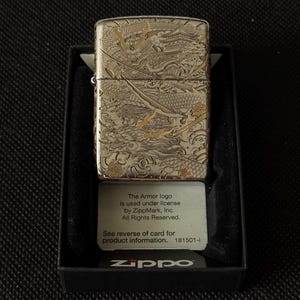May include: A silver-tone metal lighter featuring an intricate engraved design of a samurai helmet, a sword, and a dragon. The lighter is housed in a black display box with a card that reads: The Armor logo Is used under license by ZippMark, Inc.