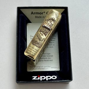May include: A gold-colored Zippo lighter with an engraved forest scene, presented in a black box. The lighter features the text "YET THE HEART THAT BEATS WITHIN IS NO LONGER MINE". The box has the Zippo logo.