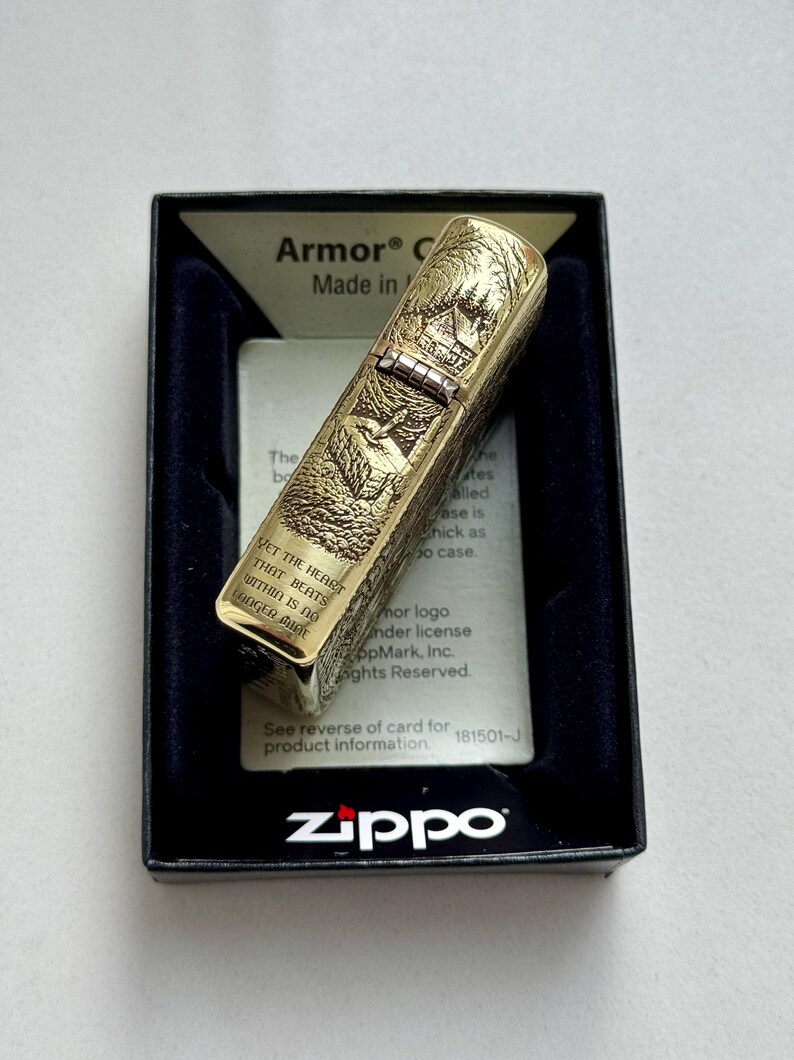 May include: A gold-colored Zippo lighter with an engraved forest scene, presented in a black box. The lighter features the text "YET THE HEART THAT BEATS WITHIN IS NO LONGER MINE". The box has the Zippo logo.