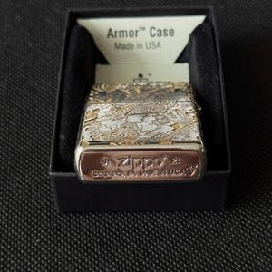 May include: A silver-tone Zippo Armor Case lighter featuring an intricate engraved design of a Japanese warrior. The base is stamped with the Zippo logo, Bradford, PA, Made in USA, and date codes. It is presented in a black box with an Armor Case insert.