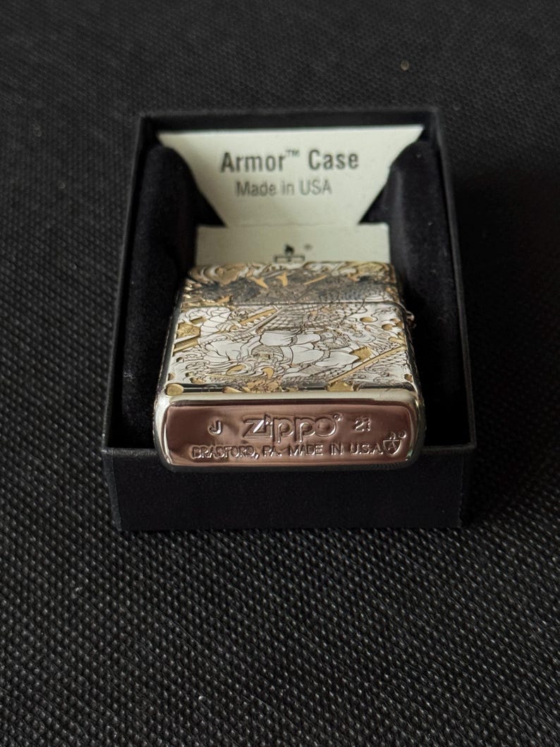 May include: A silver-tone Zippo Armor Case lighter featuring an intricate engraved design of a Japanese warrior. The base is stamped with the Zippo logo, Bradford, PA, Made in USA, and date codes. It is presented in a black box with an Armor Case insert.