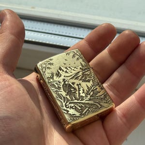 以下が含まれることがあります： 山岳風景の中で猟師と犬が描かれた、精巧な彫刻が施された長方形のゴールドトーンの金属製ライター。表面には木々、鳥、ライフルや双眼鏡などの狩猟用具が彫り込まれています。