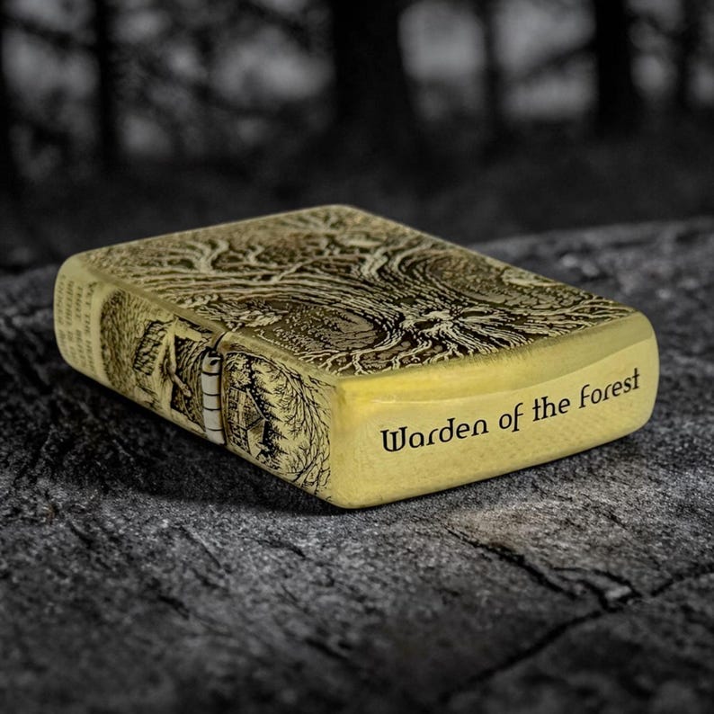 May include: A gold-colored lighter with an engraved forest scene and the words "Warden of the Forest". The lighter has a rectangular shape and a hinged lid. The forest scene includes trees and a small building.