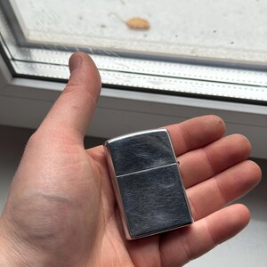 May include: A silver-colored, rectangular lighter with a brushed metal finish. The lighter is held in an open hand, with a window and foliage visible in the background.