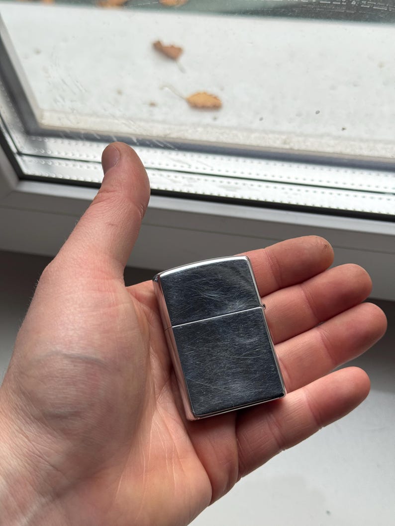 May include: A silver-colored, rectangular lighter with a brushed metal finish. The lighter is held in an open hand, with a window and foliage visible in the background.