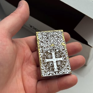 May include: A gold and silver Zippo lighter with an ornate, swirling design and the text "PAX" and other symbols. The lighter is held in a hand and is partially open, revealing the interior.