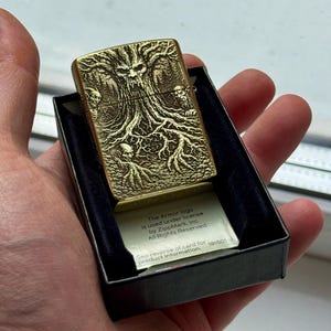 May include: A gold-colored metal lighter with a detailed tree and skull design. The lighter is in a black box with a small card that has text on it. The lighter is held in a hand.