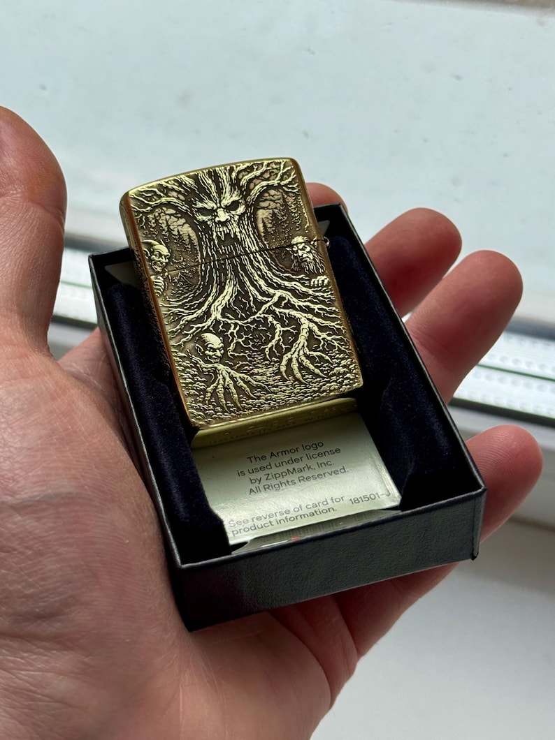 May include: A gold-colored metal lighter with a detailed tree and skull design. The lighter is in a black box with a small card that has text on it. The lighter is held in a hand.