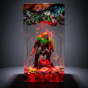 May include: A colorful resin sculpture featuring a detailed action figure of a character with a green and orange helmet, holding a gun and shield. The figure is encased in a clear rectangular block with vibrant red, yellow, and orange accents.