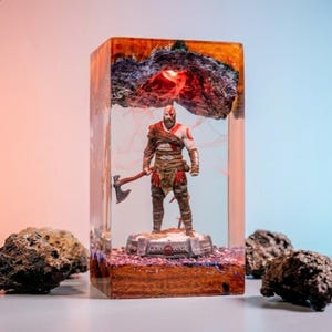 May include: A rectangular resin lamp featuring a Kratos figurine from God of War standing on a textured base. The clear resin block contains a glowing red light effect and a dark, craggy top section, with the game title printed on the base.