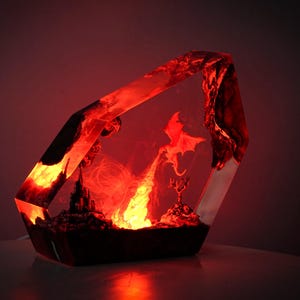 May include: A fantasy-themed resin lamp with a red glow. The lamp features a dragon, castle, and fiery elements within a geometric shape. The overall design evokes a sense of magic and adventure, perfect for a unique home decor piece.