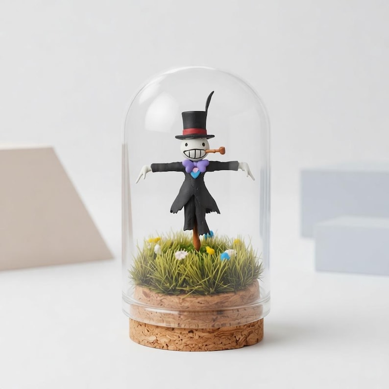 Peut inclure: Figurine fantaisiste d'un &eacute;pouvantail sous un d&ocirc;me en verre. La figurine porte un chapeau haut de forme noir, un manteau et un n&oelig;ud papillon violet. Il se tient sur un lit d'herbe verte et de petites fleurs, sur une base en li&egrave;ge.