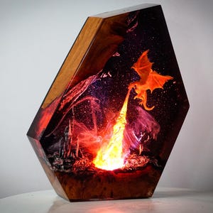 May include: A decorative resin sculpture featuring a fiery dragon breathing flames. The artwork is encased in a geometric wooden frame, with a detailed fantasy scene illuminated by an internal light source. The dragon is orange, and the flames are yellow and red.