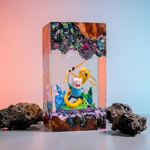 May include: A rectangular resin sculpture featuring the cartoon characters Finn and Jake from Adventure Time. The characters are set in a colorful, layered resin block with a wood grain top. The sculpture is surrounded by dark, textured rocks.