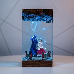 May include: A rectangular resin art piece featuring two figurines on a bridge, encased in clear resin. The top section has a wood grain design with blue swirling patterns. The figurines are dressed in blue and red attire, with a bridge and decorative elements.