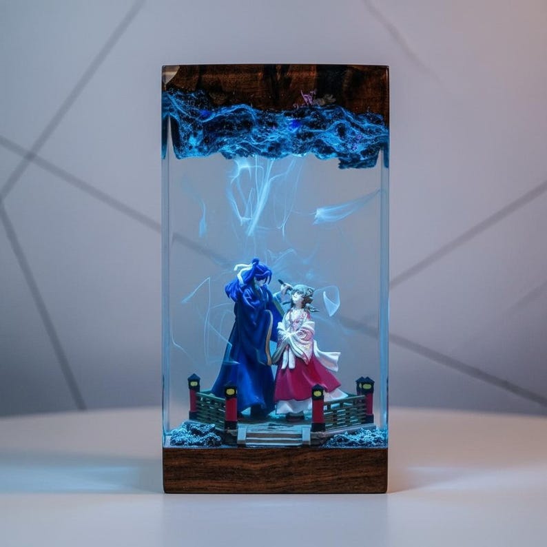 May include: A rectangular resin art piece featuring two figurines on a bridge, encased in clear resin. The top section has a wood grain design with blue swirling patterns. The figurines are dressed in blue and red attire, with a bridge and decorative elements.