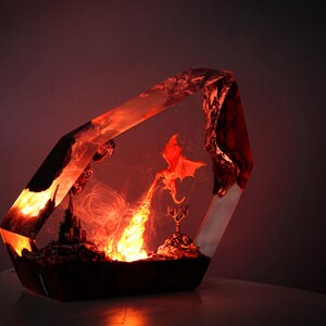 May include: A fantasy-themed resin sculpture featuring a red dragon breathing fire. The artwork is encased in a clear, geometric form, with a castle and other details illuminated by an internal light source. The overall color scheme is red and orange.