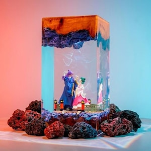 Puede incluir: Una pieza de arte de resina rectangular con dos figuras dentro de un bloque transparente. La parte superior es de color madera, con una capa texturizada azul oscuro debajo. La base está rodeada de rocas oscuras y porosas.