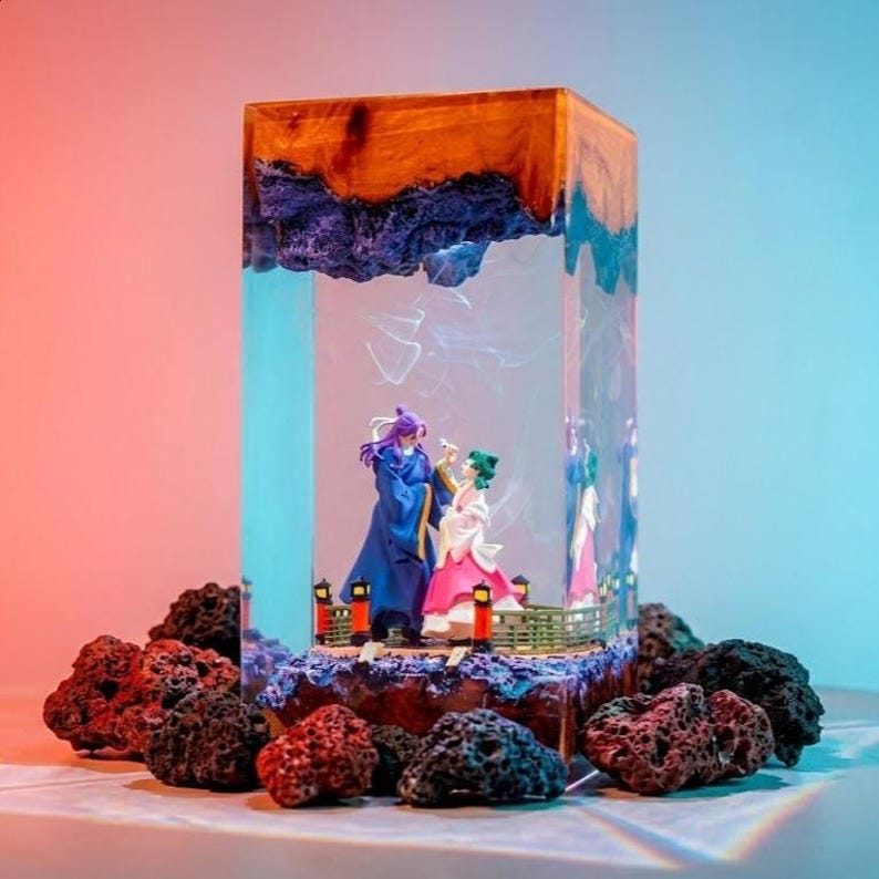 May include: A rectangular resin art piece featuring two figurines in a scene, set within a clear block. The top is wood-toned, with a dark blue, textured layer beneath. The base is surrounded by dark, porous rocks.