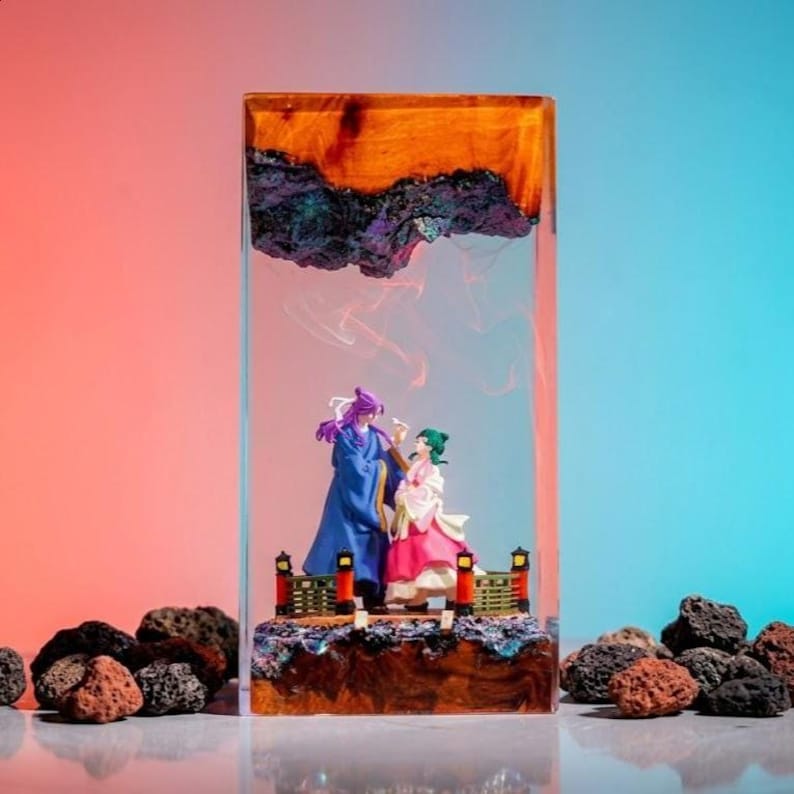 May include: A rectangular resin art piece featuring two figurines on a bridge. The resin has a layered effect with orange, purple, and black elements. The figurines are dressed in colorful traditional clothing. The background gradient shifts from pink to blue.