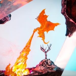 May include: An orange dragon figurine exhales a stream of fire. The dragon is perched above a rocky landscape with a small tree. The background is a gradient of white and blue.