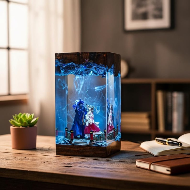 May include: A rectangular resin lamp with a wooden base and top. Inside, miniature figures stand on a bridge, illuminated by blue light. The lamp is on a wooden table, with a succulent plant in a pink pot to the left.