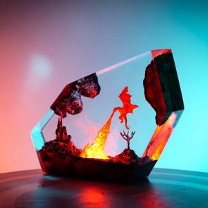 May include: A fantasy-themed resin sculpture featuring a red dragon breathing fire. The artwork is encased in a clear, geometric form, with a landscape of mountains and a barren tree. The color palette includes red, orange, and black.