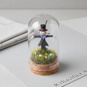 Peut inclure: Une figurine de l'&eacute;pouvantail d'un film, sous un d&ocirc;me en verre. Il porte un chapeau haut de forme noir, un costume et un n&oelig;ud papillon violet. La base est en li&egrave;ge, avec de l'herbe et des fleurs artificielles.