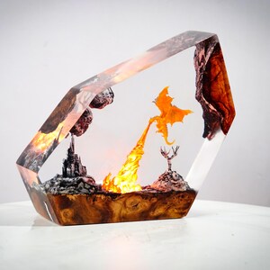 May include: A fantasy-themed resin sculpture featuring a dragon breathing fire. The artwork is encased in a clear, angled block, with a castle, trees, and a fiery landscape. The base has a wood grain appearance.