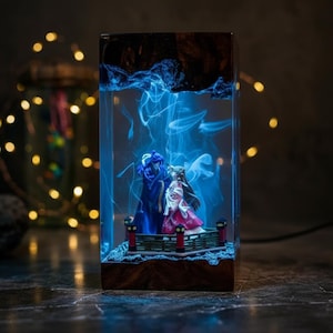 May include: A rectangular resin lamp featuring two figurines on a small bridge, illuminated by blue light. The lamp has a wooden base and a dark, textured top. The figurines are dressed in blue and pink attire.