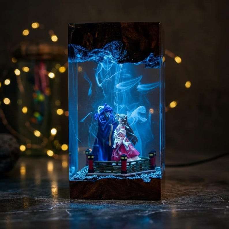 May include: A rectangular resin lamp featuring two figurines on a small bridge, illuminated by blue light. The lamp has a wooden base and a dark, textured top. The figurines are dressed in blue and pink attire.