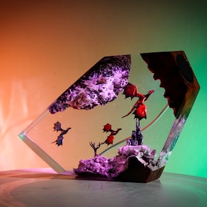 以下が含まれることがあります： 透明な樹脂製の彫刻で、赤いドラゴンが紫色の山と小さな街の上を飛んでいます。背景には、さらに小さなドラゴンが2匹飛んでいます。彫刻は木製の台座の上に置かれています。